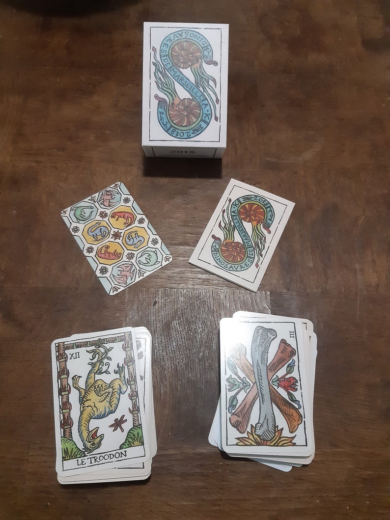 May include: A set of tarot cards with dinosaur illustrations. The cards feature colorful images of dinosaurs, including a Troodon, and the text "Le Troodon" on one card. The box for the cards is also shown, with the text "Dinosavre de Marseille AK" on the front.