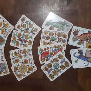 May include: A set of 10 tarot cards featuring colorful illustrations of dinosaurs and other creatures. The cards are laid out on a wooden surface. The cards are from the "Tarot of the Amazons" deck.
