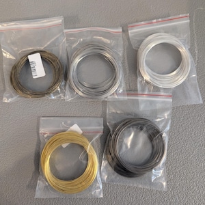 May include: Five spools of wire in clear plastic bags. The wire colors include gold, silver, black, and bronze. The spools are neatly coiled and ready for use in jewelry making or crafts.
