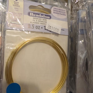 May include: A package of Beadalon memory wire for bracelets. The package includes a coil of gold-colored wire, labeled as 0.5 oz. The packaging also indicates the wire is .039 inches or 1.0 mm in diameter. The package is labeled "Large Bracelet".