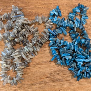 May include: Two strands of gemstone chip beads. One strand is a mix of gray and blue, the other is bright blue. The beads are irregularly shaped and strung on a thin, clear cord. The background is a light brown wooden surface.