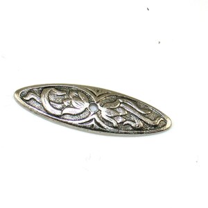 May include: Oval-shaped, silver-toned metal decorative plate with a floral design. The plate features a central hole and detailed embossed flowers and leaves. The design is symmetrical, with the floral elements arranged around the center.