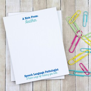 May include: A white notepad with blue text that reads "A Note From: Heather" and "Speech Language Pathologist I have ways of making you talk."  The notepad is on a woodgrain surface with colorful paper clips scattered around it.