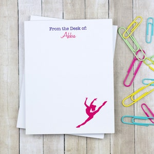 May include: A white notepad with the text "From the Desk of: Abbie" printed in purple and blue. A pink silhouette of a dancer is printed on the notepad. The notepad is surrounded by colorful paper clips.