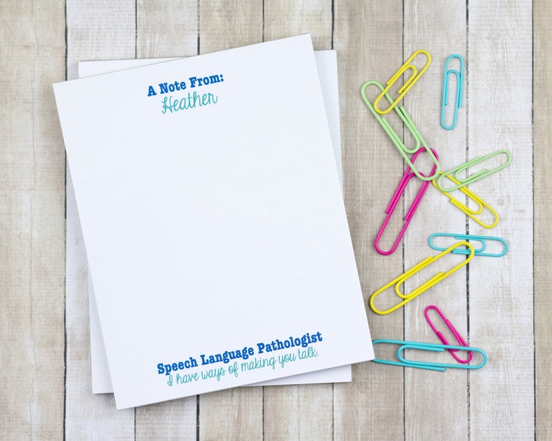 May include: A white notepad with blue text that reads "A Note From: Heather" and "Speech Language Pathologist I have ways of making you talk."  The notepad is on a woodgrain surface with colorful paper clips scattered around it.