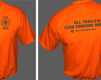 Op de afbeelding: Oranje T-shirt met een zwarte kompas en de tekst "K-9 SAR" op de voorkant. De achterkant van het shirt heeft de tekst "ALL TRAILS LEAD SOMEONE HOME. K-9 SEARCH UNIT" in zwart.