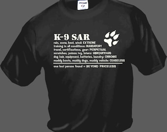 Op de afbeelding: Zwart T-shirt met witte tekst die "K-9 SAR" luidt en een beschrijving van de uitdagingen waar K-9 zoek- en reddingsteams mee te maken krijgen. De tekst bevat zinnen als "EXTREME training", "VERPLICHT reizen", "PERPETUELE certificeringen", "OVERAL voorkomende krassen", "CHRONISCHE was" en "ONOPHOUDELIJK modderig voertuig".