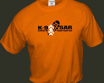 Op de afbeelding: Oranje T-shirt met een zwart-wit logo met de tekst "K-9 SAR" en daaronder de tekst "AIR SCENTING - HRD - TRACKING - TRAILING - WATER".