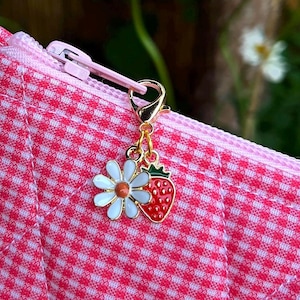 May include: A pink gingham patterned pouch with a zipper, featuring a gold charm with a white daisy and a red strawberry. The pouch is quilted and has a pink zipper. Daisies are visible in the background.