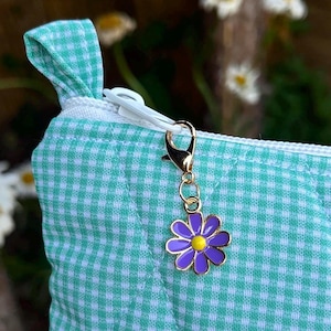 May include: Close-up of a teal and white gingham patterned pouch with a gold-colored zipper and a gold-colored charm. The charm is a purple and yellow flower. The pouch has a quilted design.