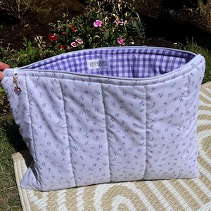 May include: A lavender and blue floral patterned quilted tote bag. The interior lining is a purple and white gingham check. The bag has a zip closure and a small flower charm. The text "PEACHHEROES" is visible on the lining.
