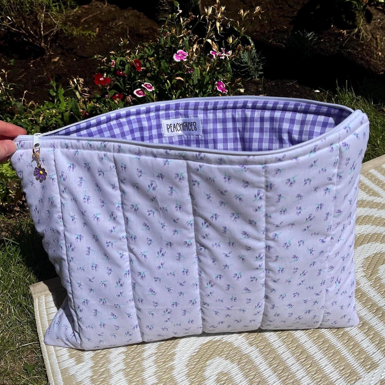 Pode incluir: Uma bolsa tote acolchoada em lavanda com um padr&atilde;o floral. O interior apresenta um forro xadrez vichy roxo e branco. A bolsa tem fecho de correr e um pequeno pendente de flor. O texto "PEACHHEROES" &eacute; vis&iacute;vel no interior.