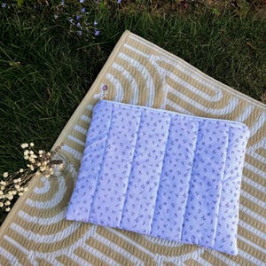 Pode incluir: Uma bolsa acolchoada azul clara com um padr&atilde;o floral, com fecho de correr. Est&aacute; sobre um tapete listrado bege e branco, com relva verde ao fundo. Um pequeno vaso de vidro com flores brancas est&aacute; &agrave; esquerda.