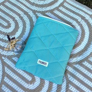 May include: A turquoise and white gingham quilted pouch with a white zipper. The pouch has a diamond pattern and a small white label that reads "PEACHFACES". A small glass vase with dried flowers sits to the left of the pouch.