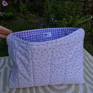 Pode incluir: Bolsa acolchoada com estampa floral em fundo branco. O forro interno &eacute; xadrez vichy roxo e branco. A bolsa tem formato retangular e o texto "PEACHFACES" &eacute; vis&iacute;vel no interior.