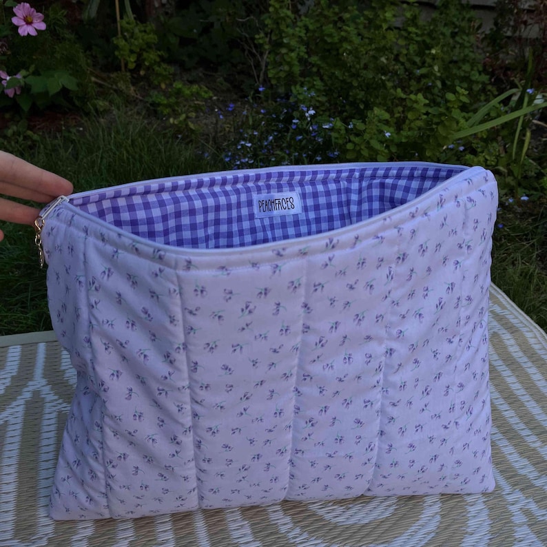 Pode incluir: Bolsa acolchoada com estampa floral em fundo branco. O forro interno &eacute; xadrez vichy roxo e branco. A bolsa tem formato retangular e o texto "PEACHFACES" &eacute; vis&iacute;vel no interior.
