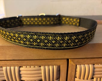 Könnte beinhalten: Schwarzes Hundehalsband mit goldenem Fleur-de-Lis-Muster. Das Halsband hat eine Metallschnalle. Das Design ist ein sich wiederholendes Muster des Fleur-de-Lis-Symbols.