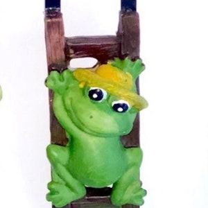 May include: A green frog wearing a yellow hat climbs a brown wooden ladder with black metal hooks. The frog has black eyes and a wide smile.