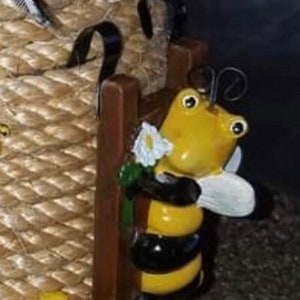 May include: A whimsical garden ornament featuring a yellow and black bee holding a white flower. The bee is positioned near a brown wooden fence and a woven basket. The bee has large eyes and antennae.