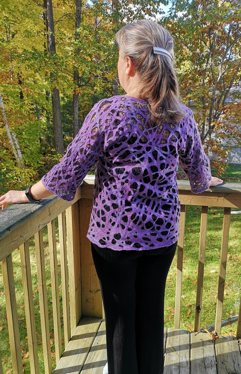 May include: A purple and black patterned sweater with a lace-like design. The sweater is worn by a person standing on a wooden deck.