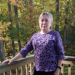 May include: A woman wearing a purple sweater with a black and white pattern stands on a wooden deck in front of a forest.
