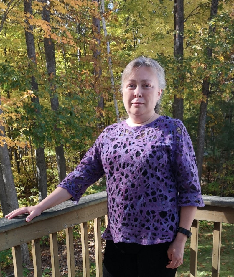 May include: A woman wearing a purple sweater with a black and white pattern stands on a wooden deck in front of a forest.