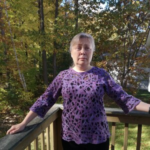 May include: A woman wearing a purple and black patterned sweater with a lace-like design. She is standing on a wooden deck with a forest background.
