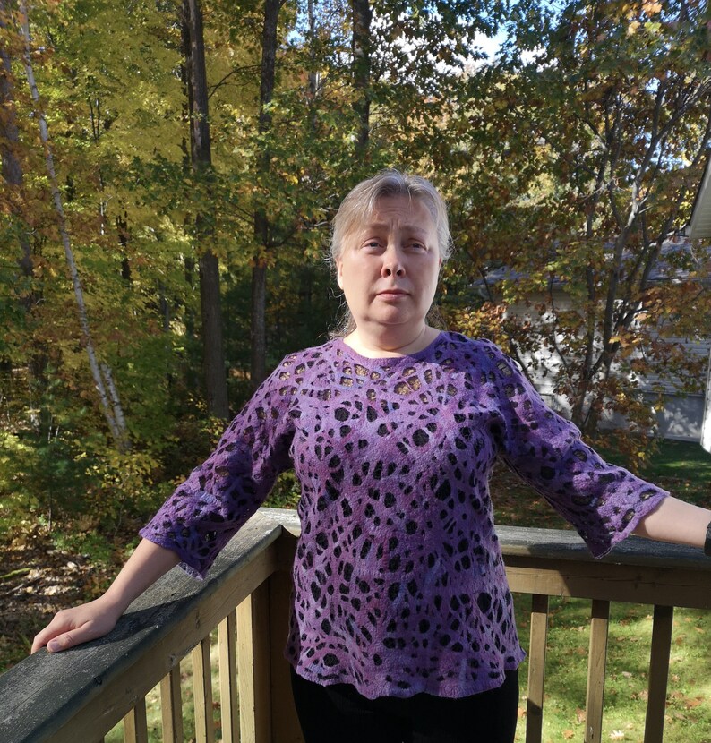 May include: A woman wearing a purple and black patterned sweater with a lace-like design. She is standing on a wooden deck with a forest background.