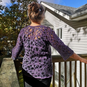 May include: A woman wearing a purple and black crocheted sweater with a lacy design. She is standing on a wooden deck with a house in the background.