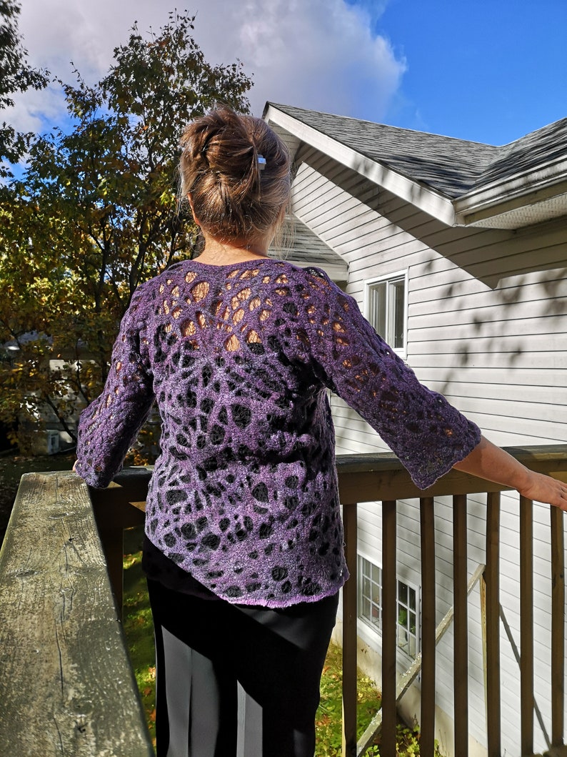 May include: A woman wearing a purple and black crocheted sweater with a lacy design. She is standing on a wooden deck with a house in the background.
