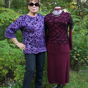 May include: A woman wearing a purple and black patterned sweater and black pants stands next to a mannequin wearing a maroon dress with a black crocheted overlay.