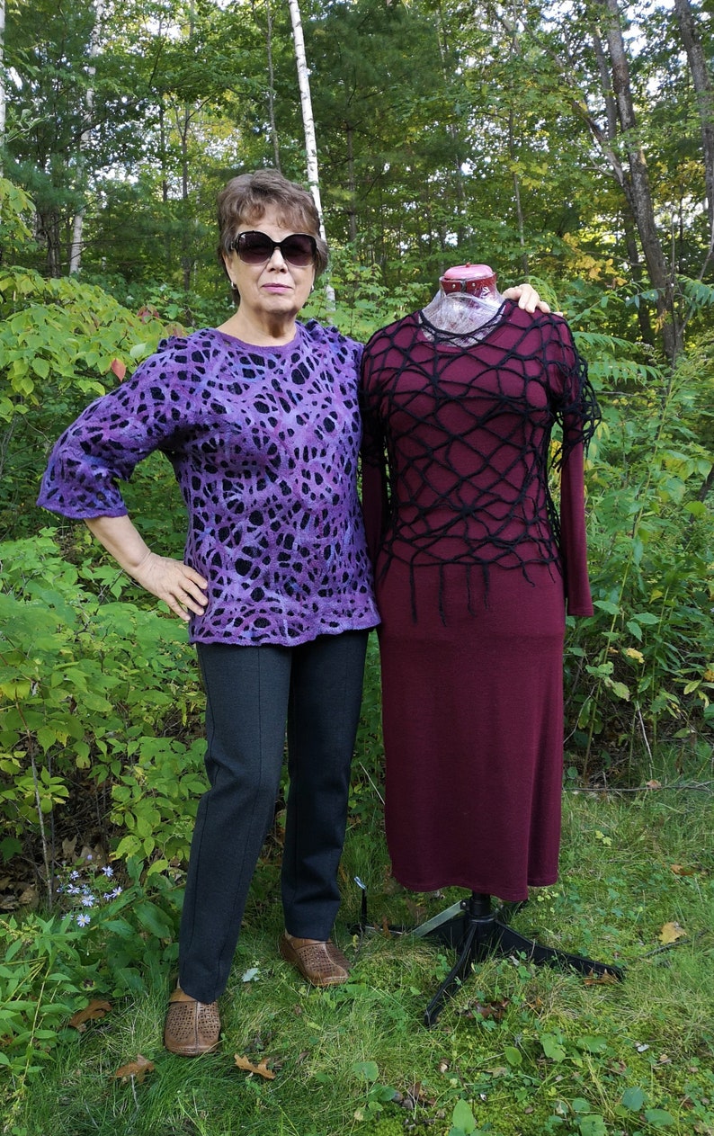 May include: A woman wearing a purple and black patterned sweater and black pants stands next to a mannequin wearing a maroon dress with a black crocheted overlay.