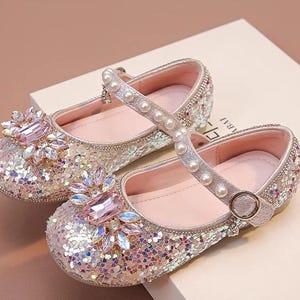May include: A pair of pink and silver glitter Mary Jane shoes. Each shoe has a strap with pearl accents and a buckle closure. The toe of each shoe is adorned with a large pink jewel and smaller clear jewels.