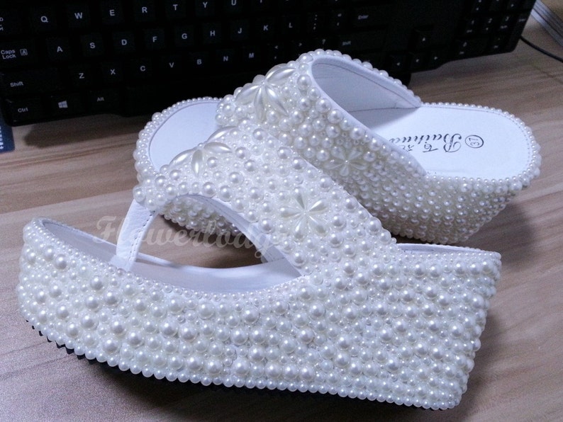 Puede incluir: Sandalias blancas de plataforma cubiertas de perlas blancas. Las sandalias tienen una suela de plataforma gruesa y una simple tira de dedo.