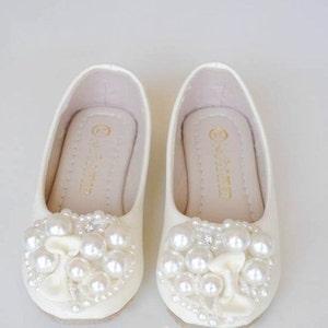 Puede incluir: Un par de zapatos de sat&eacute;n blanco con adornos de perlas. Los zapatos tienen una punta redonda y un tac&oacute;n bajo. Las perlas est&aacute;n dispuestas en un patr&oacute;n floral en la parte delantera de los zapatos.