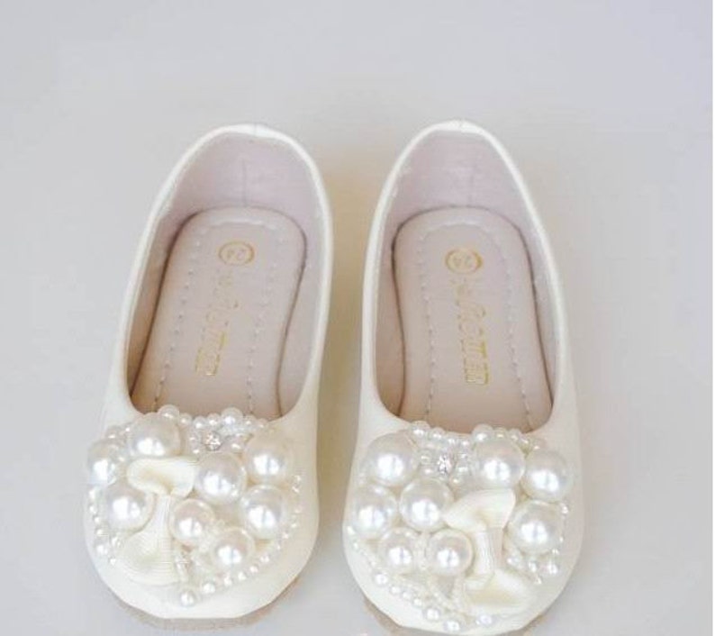 Puede incluir: Un par de zapatos de sat&eacute;n blanco con adornos de perlas. Los zapatos tienen una punta redonda y un tac&oacute;n bajo. Las perlas est&aacute;n dispuestas en un patr&oacute;n floral en la parte delantera de los zapatos.