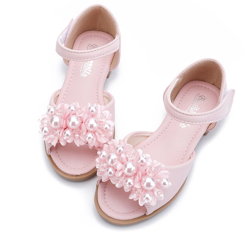 May include: Pink sandals with a pearl and flower embellishment. The sandals have an ankle strap and an open toe.