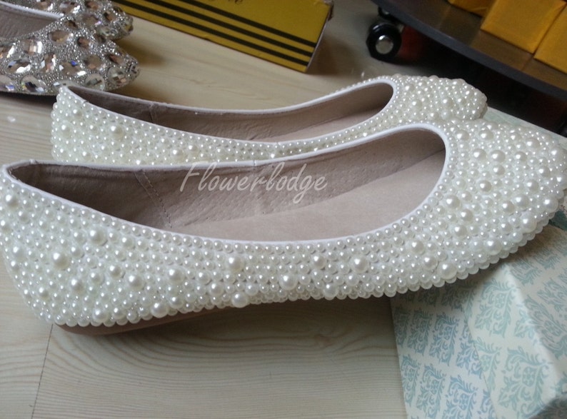 May include: A pair of white flat shoes covered in white pearl beads. The shoes are on a white surface with a patterned box in the background.