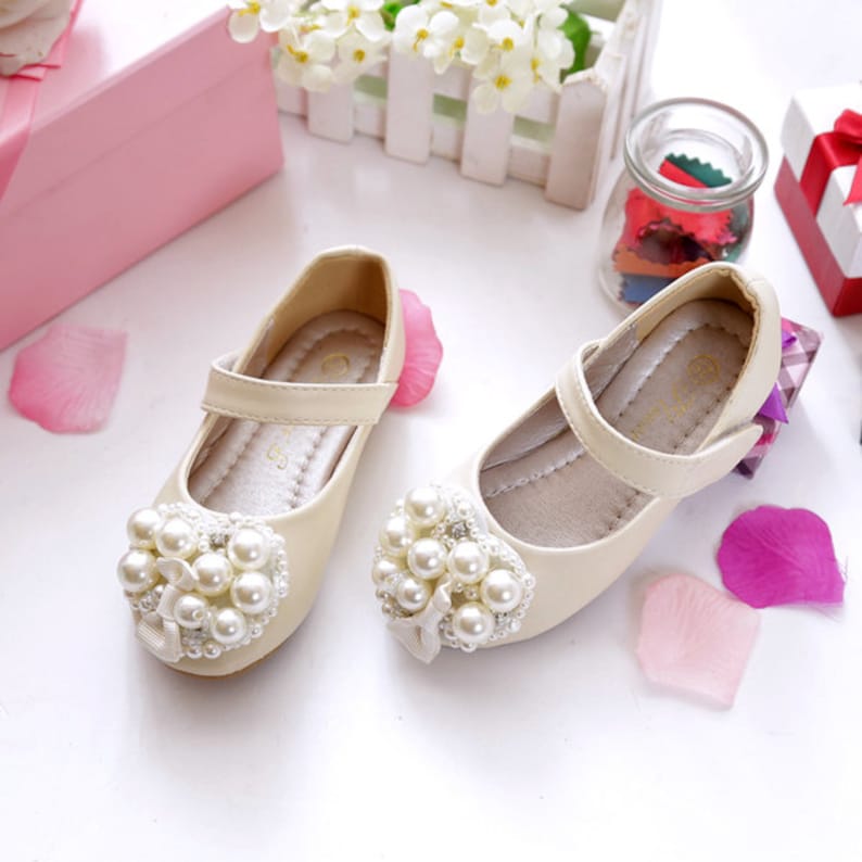 Puede incluir: Un par de zapatos de color crema para ni&ntilde;os con un adorno de perlas en la punta. Los zapatos tienen una correa sobre el pie y un cierre de hebilla.