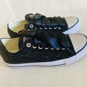 Pu&ograve; includere: Sneakers in tela nera con un nastro di raso nero legato intorno ai lacci. Le sneakers sono ricoperte di strass neri e hanno una suola in gomma bianca.