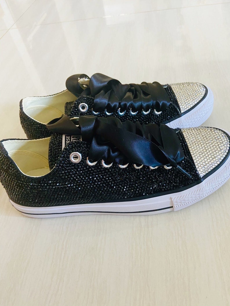 Pu&ograve; includere: Sneakers in tela nera con un nastro di raso nero legato intorno ai lacci. Le sneakers sono ricoperte di strass neri e hanno una suola in gomma bianca.