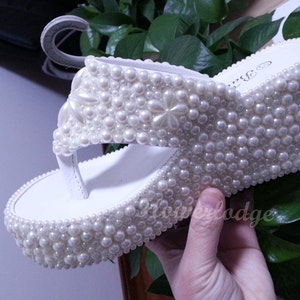 Puede incluir: Sandalias blancas de plataforma cubiertas de perlas blancas. Las sandalias tienen una suela de plataforma gruesa y una tira para el dedo del pie.