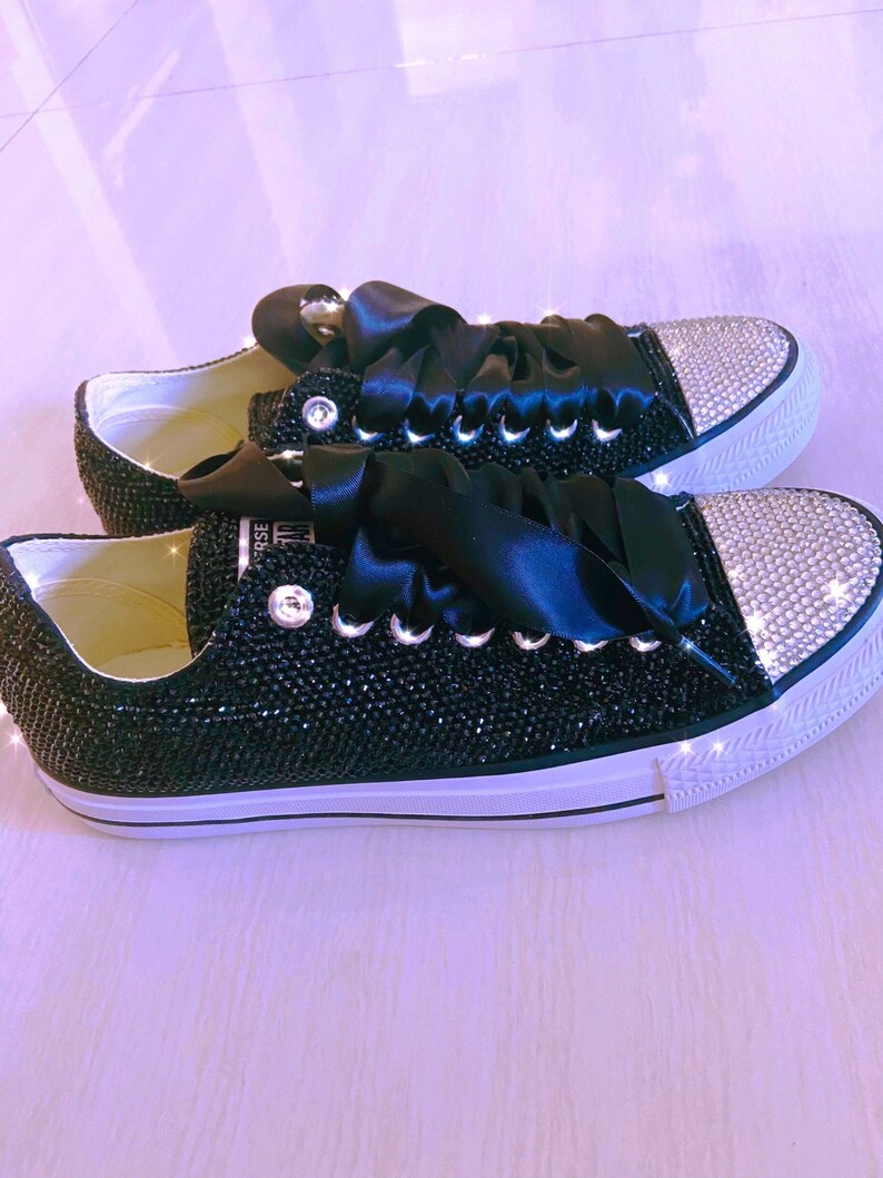 Pu&ograve; includere: Sneakers in tela nera con suola bianca e lacci in nastro di raso nero. Le sneakers sono ricoperte di strass neri e hanno un accento di strass argentato sulla punta.