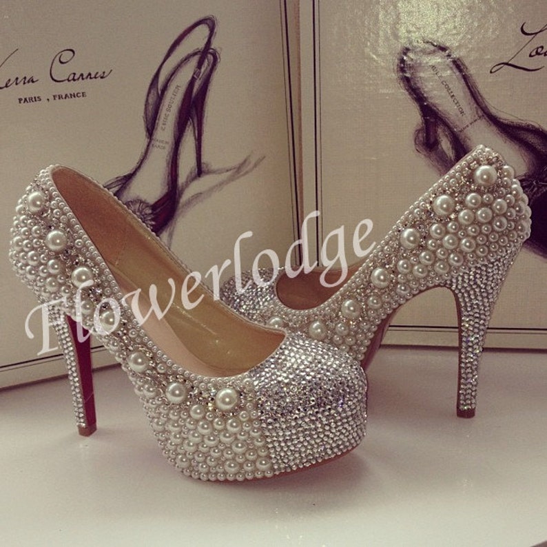May include: A pair of white high-heeled shoes with a platform sole. The shoes are covered in rhinestones and pearls.