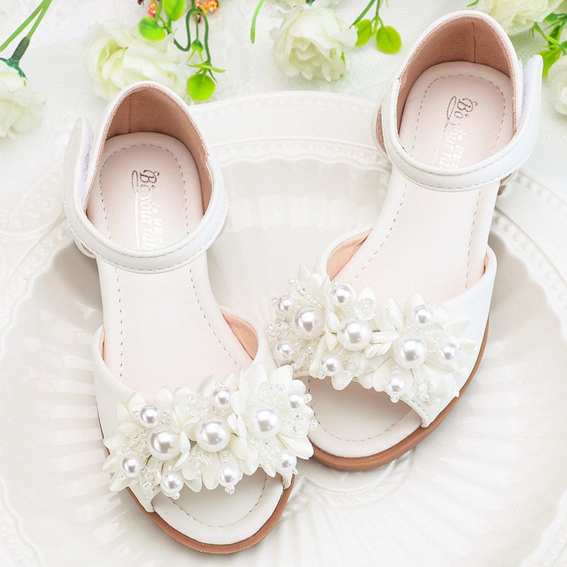 May include: White sandals with pearl and flower embellishments. The sandals have a strap around the ankle and an open toe.