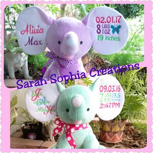 May include: Two plush elephant toys, one purple and one teal, with embroidered details. The purple elephant has the name "Alivia Max" and birth details embroidered on its ears. The teal elephant has the name "Jole Alia Hinoffa" and birth details.