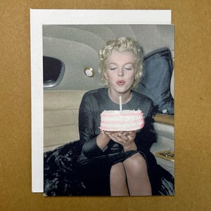 May include: A woman in a black dress blows out a candle on a pink and white layered cake. She is sitting in a car and smiling.