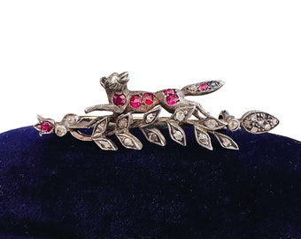 May include: A silver brooch with a fox running on a branch of leaves. The fox and leaves are decorated with red and clear gemstones.