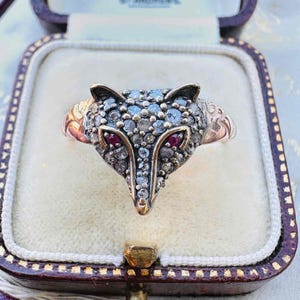 May include: A vintage fox ring with a detailed design. The fox head is encrusted with small, sparkling stones, with two red stones for eyes. The ring is set in a gold band and is displayed in a decorative box.