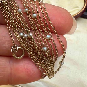 May include: Close-up of a delicate gold necklace with multiple strands. The necklace features small, lustrous pearls spaced along the chains. A gold clasp is visible, and the overall design suggests a vintage or antique style.